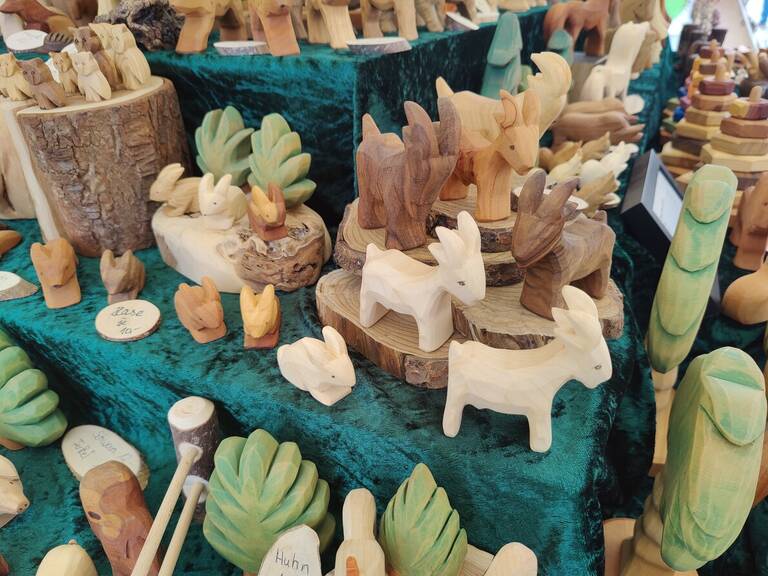 Eine Sammlung aus handgeschnitzten Holzfiguren, darunter Tiere wie Hasen und Ziegen, ist auf einem Tisch mit dunkelgrünem Stoff drapiert ausgestellt.