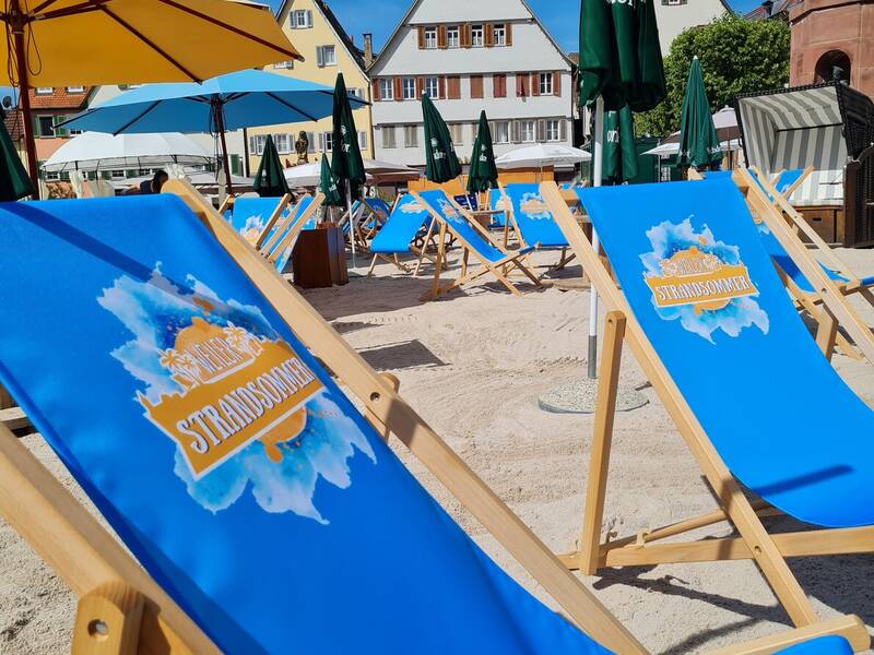 Mehrere blaue Liegestühle stehen auf Sand unter bunten Sonnenschirmen vor traditionell aussehenden Häusern mit Giebeldächern.